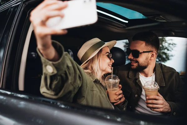 Kadın ve erkek arabada oturuyor, soğuk kahve içiyor ve akıllı telefonda selfie çekiyor, macera yolculuğunun tadını çıkarıyorlar.
