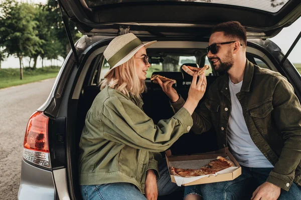 Kadın ve erkek, bir araba bagajında oturup pizza yiyor.