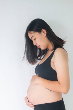Ayakta duran ve karnını beyaz arka planda tutan hamile kadın.