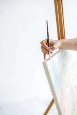 Siyah elbiseli ve önlüklü genç bir kadın parlak bir odada oturur ve resim yapar, yağlı boyaları Malbert 'le birlikte tuvale sürer.