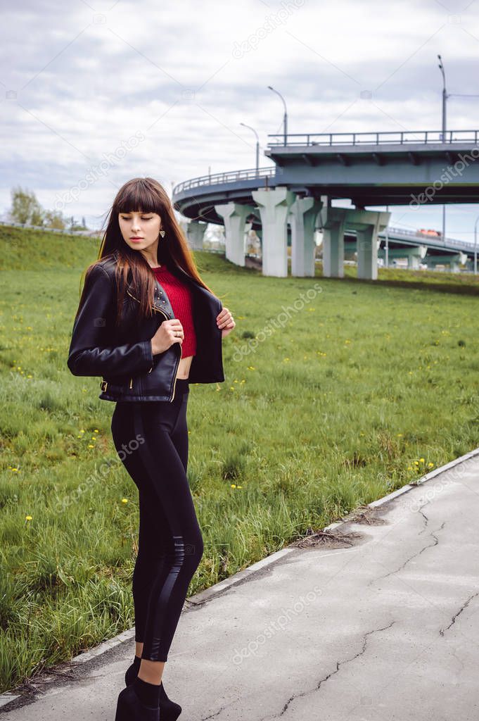 young beautiful stylish woman in fashionable black clothes, a burgundy cardigan and a dark leather jacket posing outdoors near the main road junction