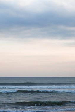 Sakin okyanus dalgalarının kıyıya doğru yuvarlanışının dikey fotoğrafı yumuşak bir gradyan gökyüzünün altında alacakaranlıkta pembeden pembeye geçiş yapan deniz ve gökyüzü arasında açık bir kontrast çizgisi ile birlikte.