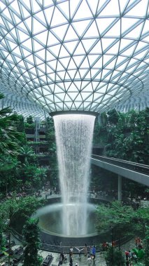 İnci, Singapur Changi Havalimanı, Singapur - 16 Ağustos 2025: dünyanın en uzun kapalı şelalesi olan Yağmur Girdabı 'nın dikey fotoğrafı, yağmur ormanları ağaçlarıyla çevrili ve yanında trenle yan yana.