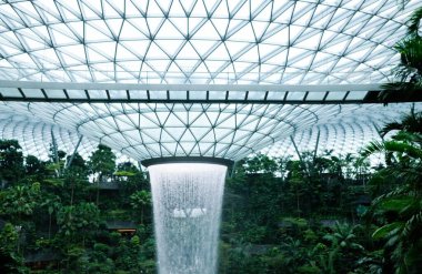 İnci, Singapur Changi Havalimanı, Singapur - 16 Ağustos 2025: dünyanın en uzun kapalı şelalesi, Yağmur Girdabı, gündüz vakti yağmur ormanları ağaçlarıyla çevrili, yan tarafında tren rayı olan.