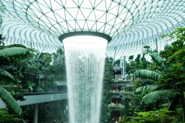 İnci, Singapur Changi Havalimanı, Singapur - 16 Ağustos 2025: dünyanın en uzun kapalı şelalesi, Yağmur Girdabı, gündüz vakti yağmur ormanları ağaçlarıyla çevrili, yan tarafında tren rayı olan.