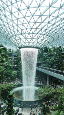 İnci, Singapur Changi Havalimanı, Singapur - 16 Ağustos 2025: dünyanın en uzun kapalı şelalesi, Yağmur Girdabı, gündüz vakti yağmur ormanları ağaçlarıyla çevrili, yan tarafında tren rayı olan.