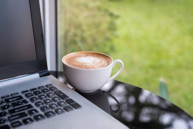 Masa üzerinde beyaz fincan sıcak kahve latte ile dizüstü bilgisayar