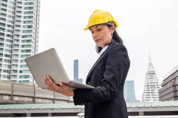 Woman engineer with yellow helmet holding laptop on urban backgr