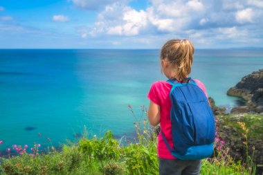 Küçük mavi sırt çantası taşıyan ve küçük bir uçurumun üzerinde duran ve Cornwall, İngiltere, İngiltere'de St. Ives yakınındaki Cornish kıyı şeridi çarpıcı hayran kırmızı t-shirt giymiş küçük kız