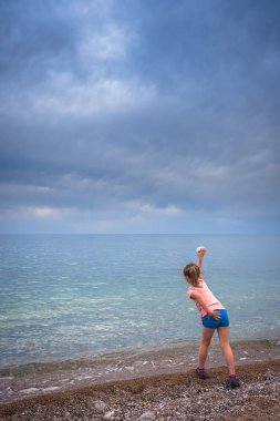 Küçük sevimli beyaz kız fırtınalı bir günde denize büyük bir taş atıyor.