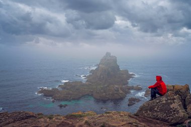 Yalnız turist bir uçurumun tepesinde oturan ve kayalık oluşumlar Lands End üzerinde-en batı noktası İngiltere, popüler turistik, Penwith Yarımadası, Penzance, Cornwall hayran su geçirmez ceket giymiş