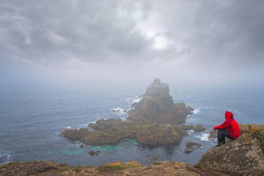 Yalnız turist bir uçurumun tepesinde oturan ve kayalık oluşumlar Lands End üzerinde-en batı noktası İngiltere, popüler turistik, Penwith Yarımadası, Penzance, Cornwall hayran su geçirmez ceket giymiş