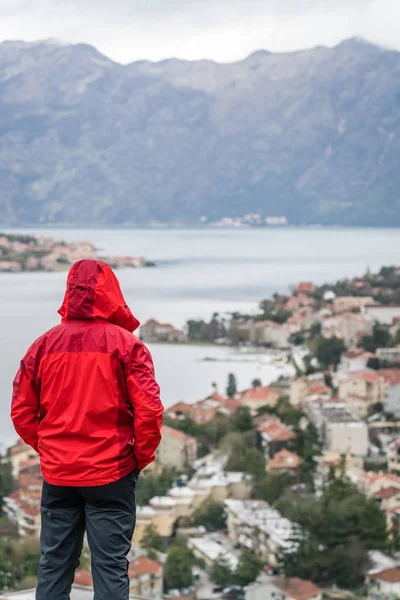 Man dressed in a red waterproof jacket standing on a lookout and looking at the view of the Kotor Bay and Old Town houses from above, Montenegro