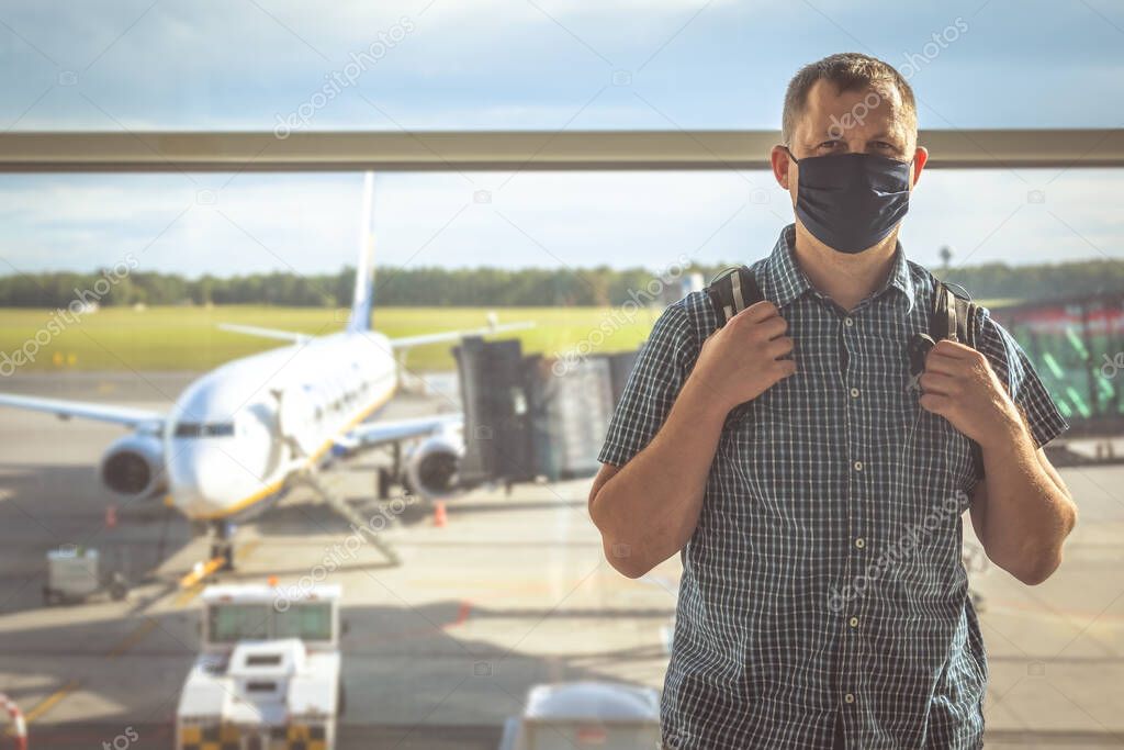 Middle aged male man with a backpack, wearing protective face mask, standing in front of large window with a view of the plane boarding shaft, traveling in the time of covid19 pandemic