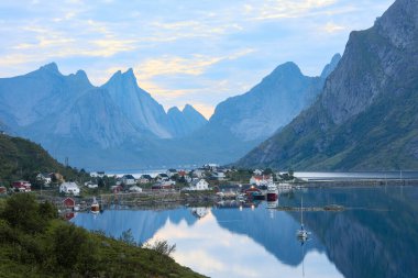 Suyun üzerinde yer alan balıkçı köyü okyanusa yansıyor yüksek dağ sıraları arasında, fiyort, pürüzsüz mavi su yüzeyi ve beyaz gecelerde Lofoten, Ramberg, Reine Norveç adasında tekneler.