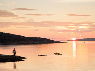 Bir deniz feneri, üç kano ve ufukta batan beyaz bir güneş, yaz mevsiminde Lofoten Adaları 'ndaki Norveç fiyortlarında güneş....