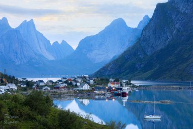 Suyun üzerinde yer alan balıkçı köyü okyanusa yansıyor yüksek dağ sıraları arasında, fiyort, pürüzsüz mavi su yüzeyi ve beyaz gecelerde Lofoten, Ramberg, Reine Norveç adasında tekneler.