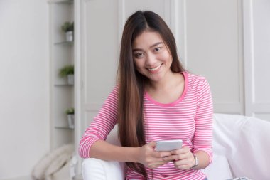 Asyalı kadın gülümseyerek ve beyaz yatak odası, arka plan bulanık smartphone kullanarak.