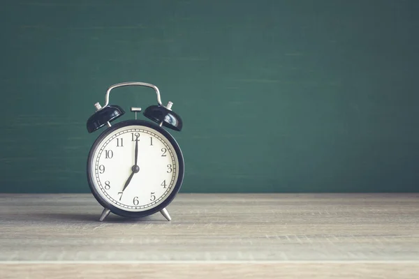 Relógio despertador na mesa de madeira no fundo do quadro-negro em sala de aula . — Fotografia de Stock