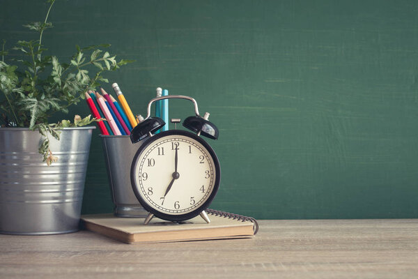 Alarm clock on wooden table on blackboard background in classroom.