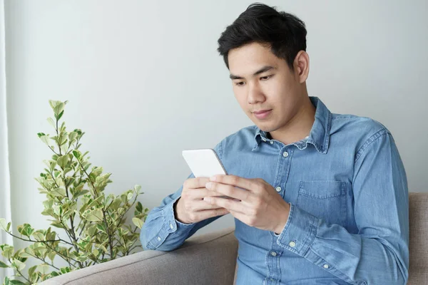 Ritratto di giovane uomo che indossa camicia blu sta guardando con smartphone e seduto al suo divano in ufficio . — Foto Stock