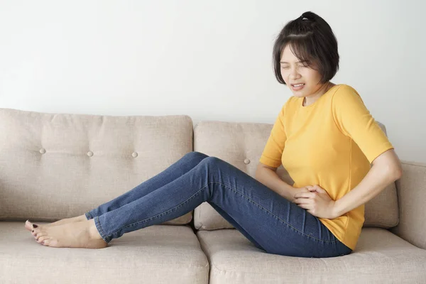 若いアジア女性の居間のソファーに座っています。彼女は痛みを伴う腹痛を起こしてください。. — ストック写真