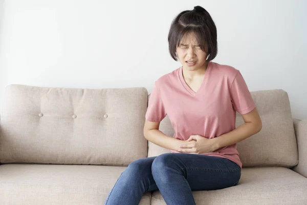 Ung asiatisk kvinna sitta på soffan i vardagsrummet. Hon har smärtsamma magont. — Stockfoto