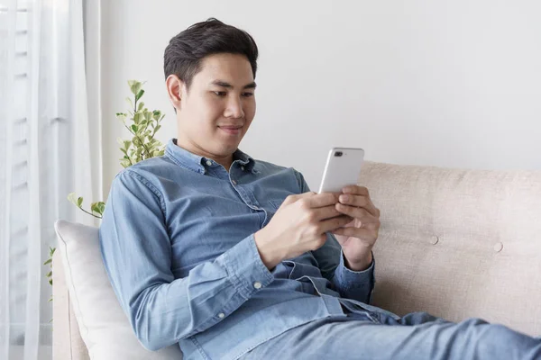 Ritratto di giovane uomo che indossa camicia blu sta guardando con smartphone e seduto al suo divano in ufficio . — Foto Stock