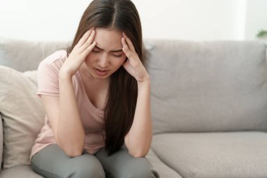 Asyalı kadın evde kanepede ağrı içinde kafasına ellerini tutuyor