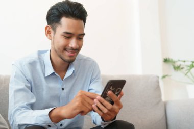 Mavi gömlek giyen genç Asyalı yakışıklı portresi akıllı telefon kullanarak okuyor ve evde oturma odasında kanepede oturuyor