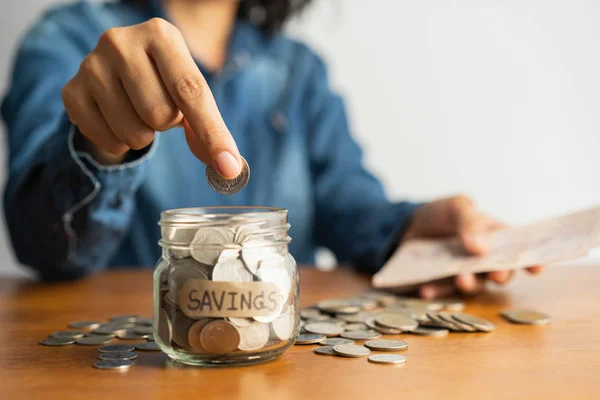 La mano de la mujer está poniendo una moneda en una botella de vidrio y una pila de monedas en una mesa de madera marrón — Foto de Stock