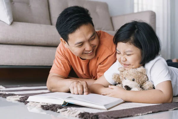 Asian father and daughter are reading book while lying on the carpet in the living room at her home.