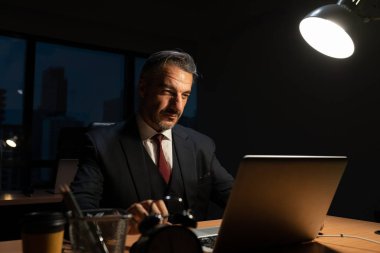 Üst düzey erkek işadamı ofis masasında dizüstü bilgisayarla çalışırken geceleri ofiste fazla mesai yapıyor.