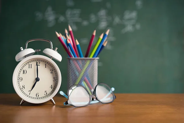Συναγερμός ρολόι λευκό και πολύχρωμο μολύβι, Γυαλιά τοποθετήστε στο ξύλινο τραπέζι στον μαυροπίνακα φόντο στην τάξη — Φωτογραφία Αρχείου