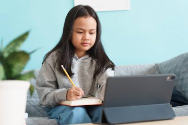 Asyalı küçük kız evdeki oturma odasında kanepede otururken internet üzerinden tablet üzerinde ders çalışıyor. Asyalı çocuklar ev ödevi izler ve yazar. Evde çevrimiçi öğrenim kavramı