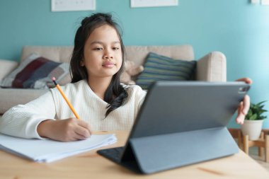 Eğitim. Asyalı kız, evde dijital tabletten internet üzerinden özel ders öğreniyor. Asyalı çocuklar bilgisayar izler ve ödev yaparlar. Evde çevrimiçi öğrenim kavramı