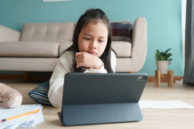 Eğitim. Asyalı kız, internet üzerinden sabah dijital tabletten özel ders alıyor. Asyalı çocuklar yerde yatarken bilgisayarla ödev yapıyorlar. Evde çevrimiçi öğrenim kavramı