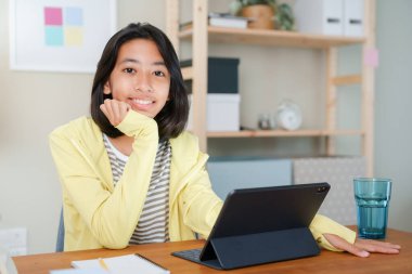 Asyalı kızın portresi internet üzerinden tablet, Asya 'daki çocuk mutluluğu üzerinden çalışıyor ve geceleri masada otururken güvenle kameraya bakarak gülümsüyor, evde online öğrenim kavramı