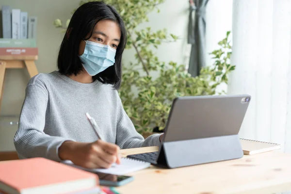 Asian woman wear medical mask while sitting  learning online with laptop computer at home, concept online learning at home