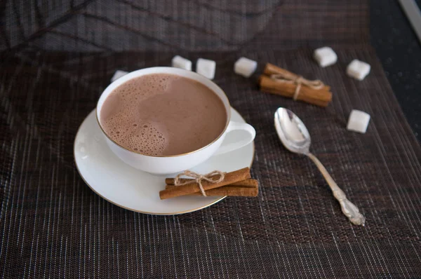 An elegant white cup of cocoa with some cinnamon sticks and sugar cubes around it. There is a teaspoon next to the cup.