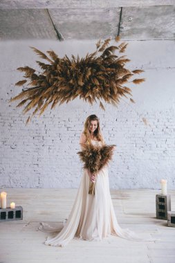 bej bir gelinlik elleri ve saman kompozisyon eski duvarın ve mum arka planda başının üstünde saman sheaves ile gülümseyen sevgili gelin. Düğün anlayışı. Studio fotoğraf