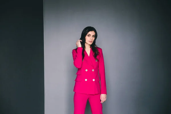 young stylish woman in pink suit posing in dark room with grey walls