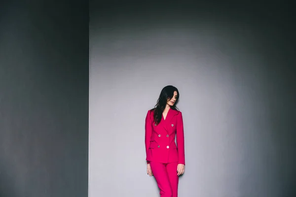 young stylish woman in pink suit posing in dark room with grey walls