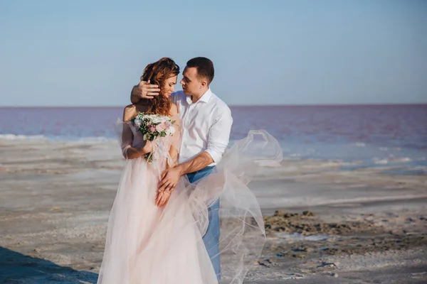 Belos recém-casados na costa do mar e noivo gentil, tocando o cabelo das noivas. vista frontal . — Fotografia de Stock