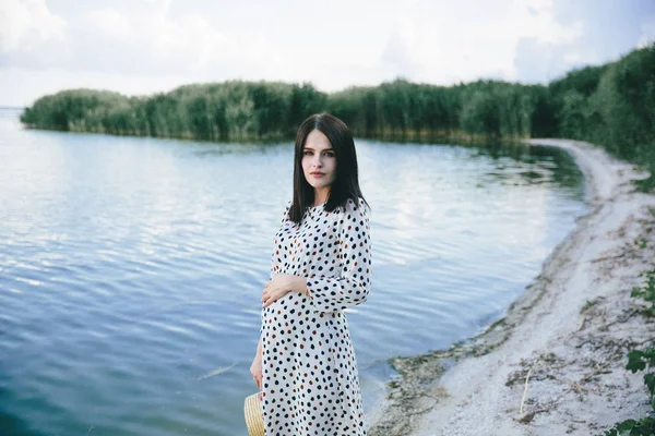 Pregnant woman in polka dot dress standing on the river background and touching her belly — Stock Photo, Image