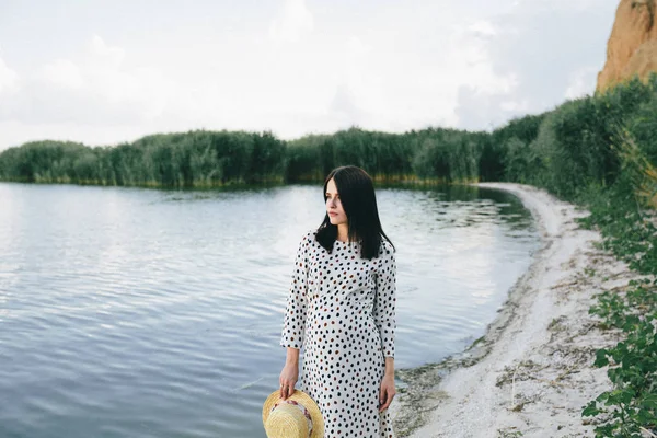 Pregnant woman in polka dot dress standing on the river background and touching her belly — Stock Photo, Image