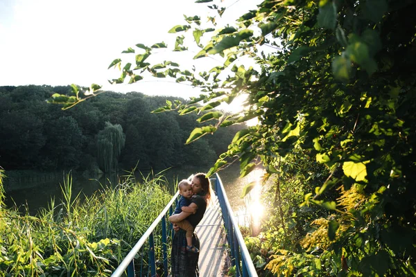 Vista frontal madre e hijo bebé abrazándose en el puente en el lago en la hora de la tarde de verano —  Fotos de Stock