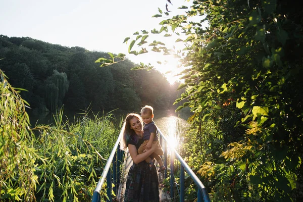 Front view mother and baby son hugging on the bridge on lake in summer evening time Stock Photo