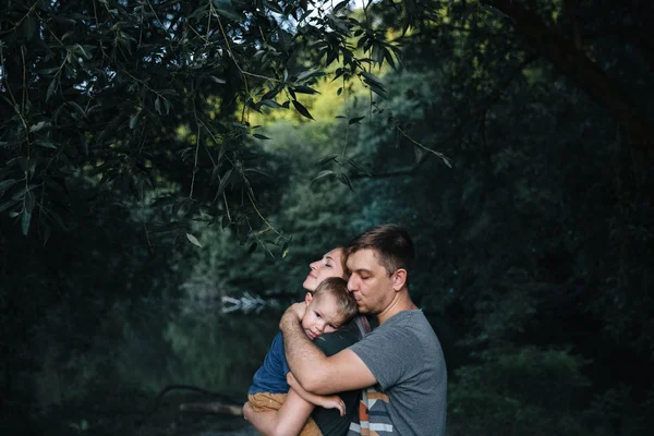 Счастливая молодая семья отец, мать и маленький сын обнимаются и трогают на улице, играя вместе в летнем парке Лицензионные Стоковые Изображения