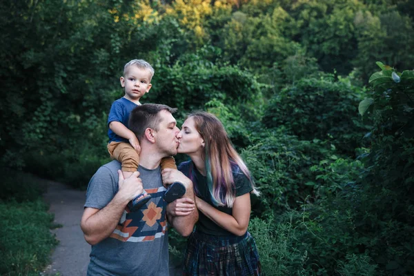พ่อแม่และลูกชายที่มีความสุข กอดและจูบกันข้างนอก เล่นด้วยกันในสวนฤดูร้อน รูปภาพสต็อก